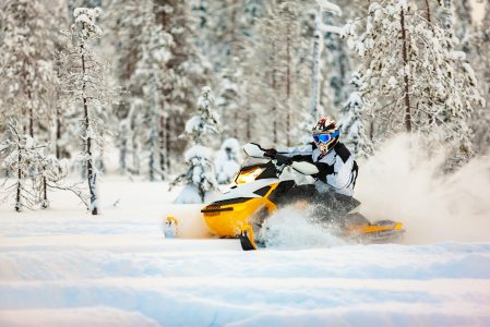 The rider in gear with a helmet drifting on a snowmobile on a deep snow surface on a background of snowy landscaping nature and winter forest.